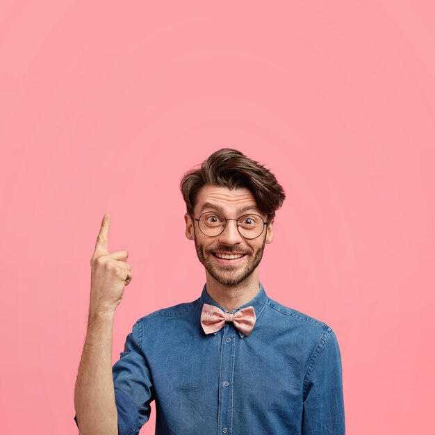 Positiver kaukasischer Mann mit trendiger Frisur, dunkler Borste, froh, etwas oben zu bemerken, zeigt mit dem Zeigefinger