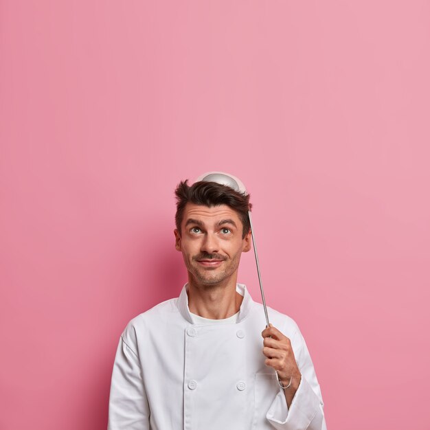 Positiver männlicher Koch posiert mit Kelle auf dem Kopf, bereitet Suppe zu, trägt weiße Uniform, hält Küchenutensilien, macht Restaurantküche, schaut oben