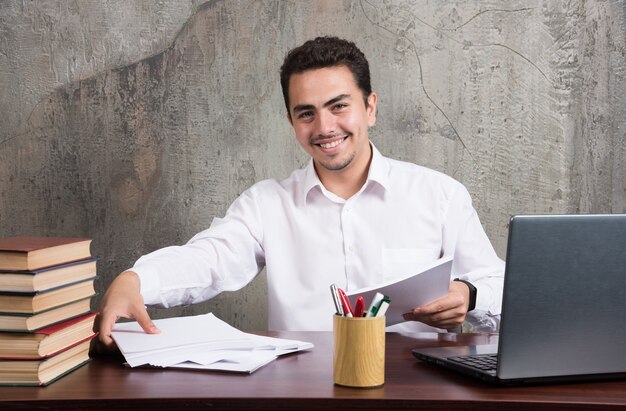 Positiver Mann, der Blätter des Papiers hält und am Schreibtisch sitzt. Hochwertiges Foto