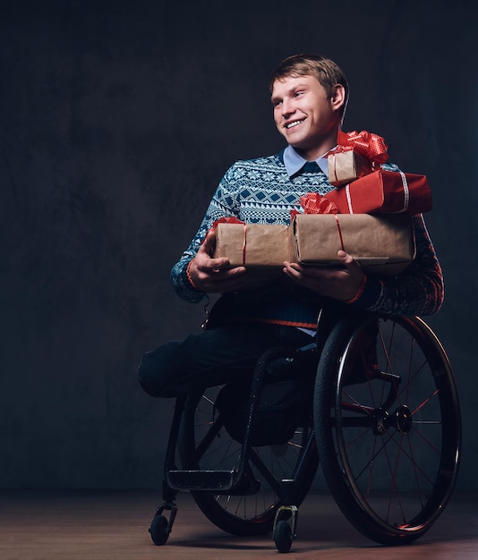Kostenloses Foto positiver mann im rollstuhl mit weihnachtsgeschenken über dunkelgrauem hintergrund.
