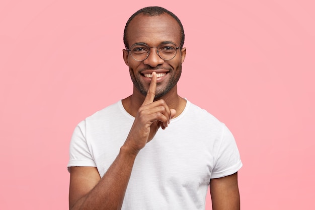 Positiver Mann macht leise Geste, hat weiße Zähne, bittet darum, leise zu sein und keinen Lärm zu machen, steht allein drinnen, trägt lässiges weißes T-Shirt