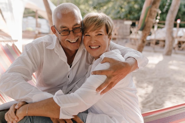 Positiver Mann mit grauem Schnurrbart in Brille und weißem Hemd lacht und umarmt seine Frau mit blonden Haaren in Loght-Bluse am Strand