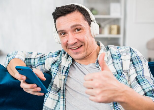 Positiver Mann mit herauf hörender Musik des Daumens in den Kopfhörern und auf Smartphone auf Sofa surfen