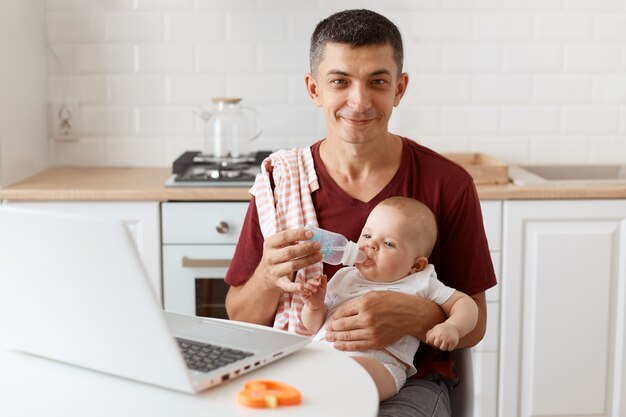 Positiver Mann mit Lächeln, der ein burgunderfarbenes Freizeit-T-Shirt mit Handtuch auf der Schulter trägt, sich um das Baby kümmert und von zu Hause aus online arbeitet, seiner Tochter Wasser gibt, schaut in die Kamera.