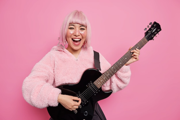 Kostenloses Foto positiver modischer weiblicher rockstar mit rosa frisur spielt akustikgitarre hat eine eigene musikband, die in einem stilvollen mantel gekleidet ist und einen neuen song für ihr album posiert drinnen. glückliche stilvolle gitarristin