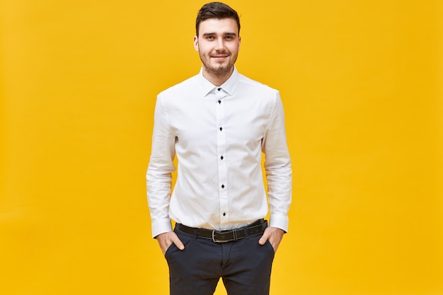 Positiver selbstbewusster junger kaukasischer männlicher Büroangestellter, der weißes formelles Hemd und klassische Hose mit Gürtel trägt, glücklichen Gesichtsausdruck hat, Hände in den Taschen hält und freudig lächelt