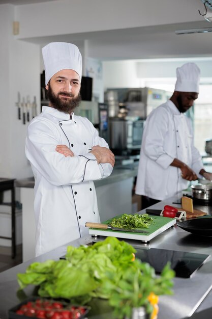 Positiver Sous-Chef, der frische Kräuter schneidet, die in Gourmetgerichten verwendet werden, während er in die Kamera schaut. Gastronomieexperte in Kochuniform beim Vorbereiten der Garnitur für das Abendessen.