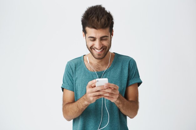 Positiver spanischer bärtiger Mann in stilvollem blauem T-Shirt, guter Frisur und Bart, Musik mit Kopfhörern hörend und durch soziale Netzwerke auf seinem Handy schauend.