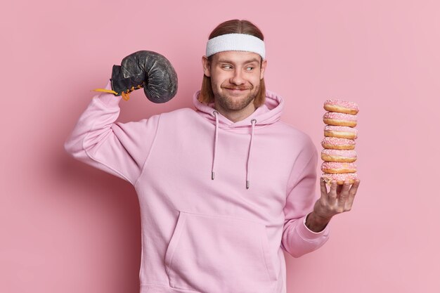 Positiver Sportler hebt Arm zeigt Bizeps nach dem Training trägt Boxhandschuh hält Haufen köstlicher Donuts in Sweatshirt Stirnband gekleidet.