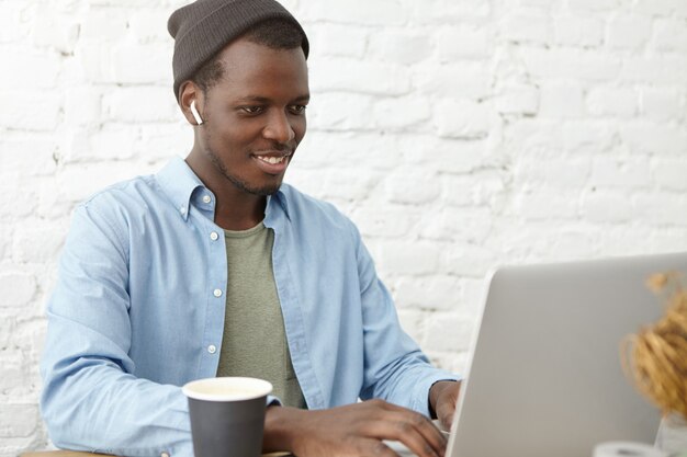 Positiver stilvoller junger afroamerikanischer Texter im Hut, der drahtlose Ohrhörer trägt, die mit seinem Chef über Videokonferenzanruf auf einem allgemeinen Laptop-Computer plaudern, auf Bildschirm schauen und fröhlich lächeln