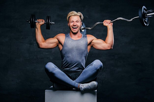 Positiver und lächelnder blonder sportlicher Mann ist in grauer Sportbekleidung gekleidet, sitzt auf einer Kiste und hält Barben.