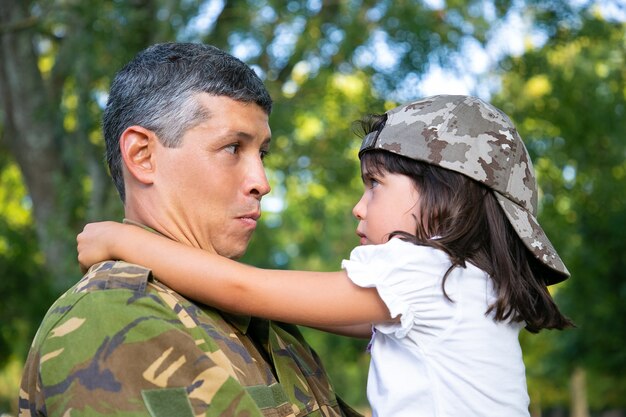 Positiver Vater in Tarnuniform, die beleidigte Tochter in den Armen hält und Mädchen im Freien umarmt, nachdem er von der militärischen Missionsreise zurückgekehrt ist. Nahaufnahme. Familientreffen oder Rückkehr nach Hause Konzept