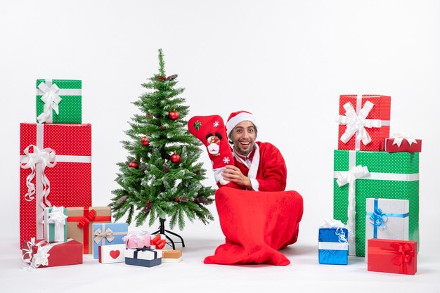 Positiver Weihnachtsmann, der auf dem Boden sitzt und Weihnachtssocke nahe Geschenken und geschmücktem Neujahrsbaum auf weißem Hintergrund trägt