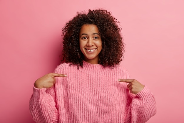 Positives Afro-Mädchen zeigt auf sich selbst, zeigt auf die Brust, sieht prahlerisch aus, trägt einen übergroßen Strickpullover, teilt gute Nachrichten mit, lächelt angenehm, isoliert über der rosa Wand