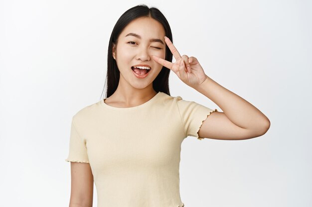 Positives asiatisches Mädchen, das ein Friedenszeichen zeigt, das zwinkert und die Zunge glücklich im gelben T-Shirt auf weißem Hintergrund hält. Kopieren Sie Platz