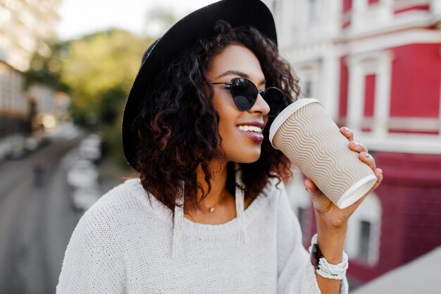 Positives Bild im Freien der lächelnden hübschen schwarzen Frau im weißen Pullover und im schwarzen Hut, die Kaffee genießen, um zu gehen. Städtischer Hintergrund.