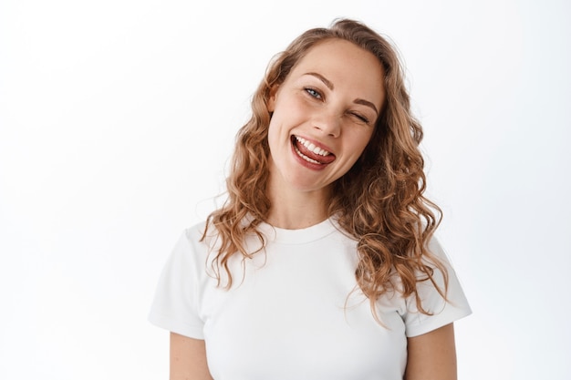 Positives blondes Mädchen, das zwinkert, Zunge zeigt und glücklich lächelt, optimistisch bleibt, mit Freude nach vorne schaut, im T-Shirt gegen weiße Wand steht