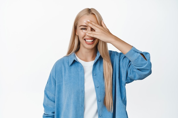 Positives blondes mädchen lacht und lächelt, späht durch die finger, offene augen, steht auf weiß