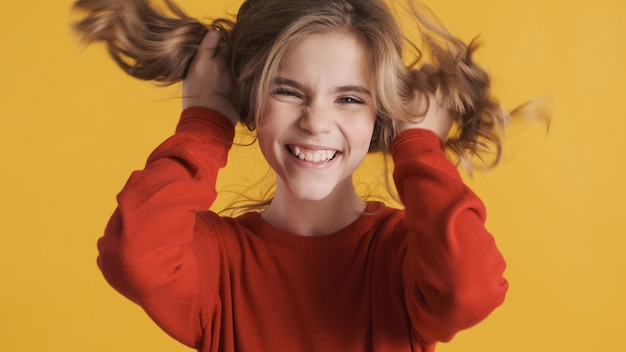 Positives blondes Teenager-Mädchen in rotem Pullover, das mit Haaren spielt, die auf gelbem Hintergrund herumalbern