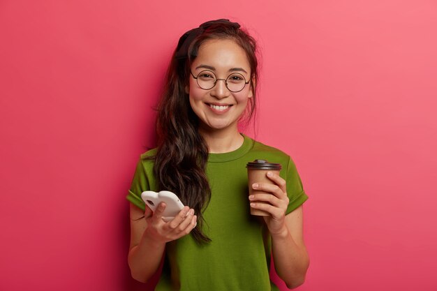 Positives entzückendes ethnisches Mädchen scrollt Fotos im Smartphone, verwendet modernes Smartphone und trinkt Kaffee zum Mitnehmen, fühlt sich berührt und entzückt, trägt runde Brillen, verwendet Einkaufswebsite