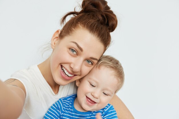 Positives Familien-Selfie mit junger Mama und zahnlosem Kind, die zusammen auf weißer Wand lächeln. Der spielerische Geisteszustand und die fröhliche Stimmung einer attraktiven Frau machen diesen Schuss fabelhaft und herzerwärmend.