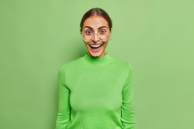 Positives frohes europäisches weibliches Model ist überrascht, kichert glücklich, trägt eine transparente Brille und einen Rollkragenpullover gegen eine leuchtend grüne Wand