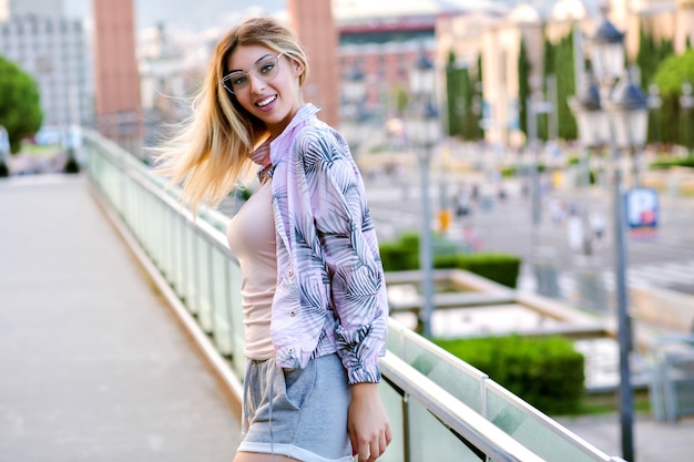 Positives Frühlingsporträt der glücklichen blonden stilvollen Frau im Freien, das trendige Kleidung für Sportfitness und -lauf trägt und am europäischen Stadtplatz, weiche Pastellfarben aufwirft.