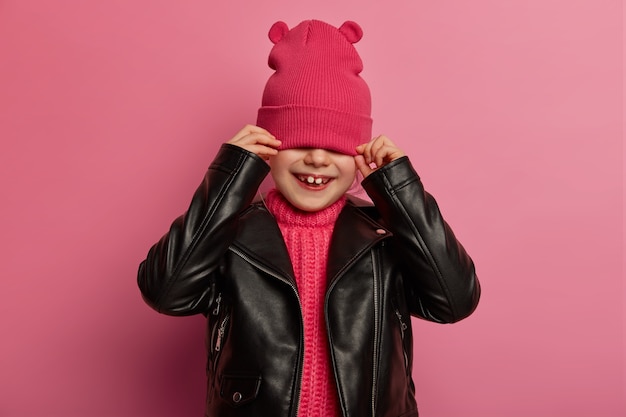 Kostenloses Foto positives kleines kind versteckt gesicht mit rosa hut, bedeckt augen, trägt lederjacke, hat verspieltes fröhliches lächeln, posiert gegen rosige wand, fühlt sich optimistisch, probiert modisches outfit an. kinderkonzept