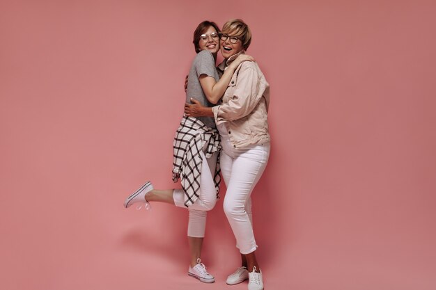 Positives Mädchen im grauen T-Shirt und im karierten Hemd lächelnd und umarmend moderne Frau mit blondem Haar in weißen Hosen und beige Jacke auf rosa Hintergrund.