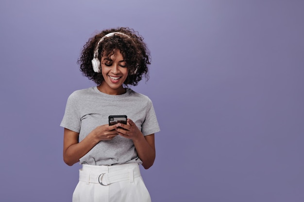 Positives mädchen in hellem t-shirt mit einem lächeln, das am telefon chattet und musik hört stilvolle frau in grauem t-shirt und weißen shorts, die lieder in kopfhörern genießen