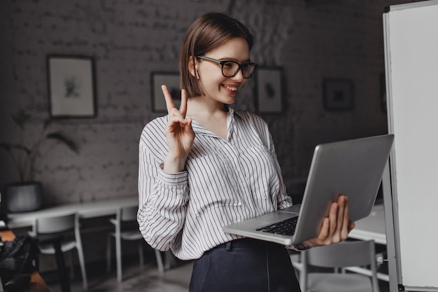 Positives Mädchen in Kopfhörern und Brille zeigt Friedenszeichen, das auf Video im Laptop am Arbeitsplatz spricht.