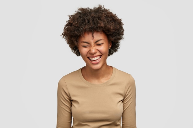 Positives menschliches Gefühlskonzept. Glückliche lockige junge Afroamerikanerin mit fröhlichem Ausdruck, kichert und lacht über etwas Lustiges, blinzelt Gesicht vor Glück, isoliert über weißer Wand