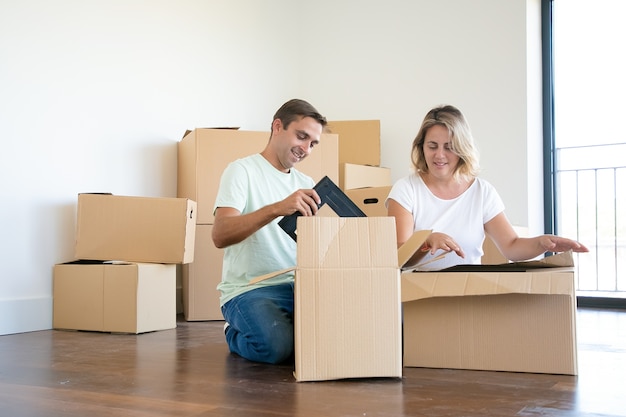 Positives Paar, das Dinge in der neuen Wohnung auspackt, auf dem Boden sitzt und Gegenstände aus offenen Kisten nimmt