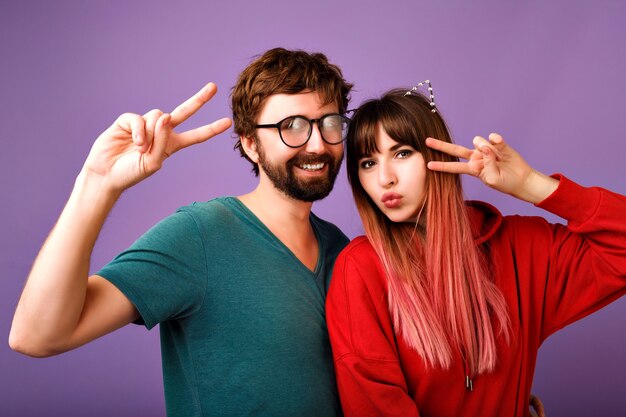 Positives Porträt des verrückten jungen Hipster-Paares, das Spaß zusammen hat, Friedensgeste zeigt, die lacht und schreit, stilvolles lässiges Kapuzenpulli und T-Shirt, Familien- und Freundbild trägt