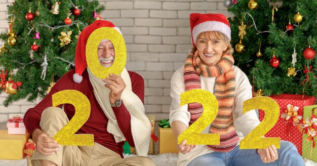 Positives seniorenpaar, das die kamera mit einem lächeln anschaut, das die goldene nummer 2022 gegen die mauer und die weihnachtsbäume während der neujahrsfeier zeigt