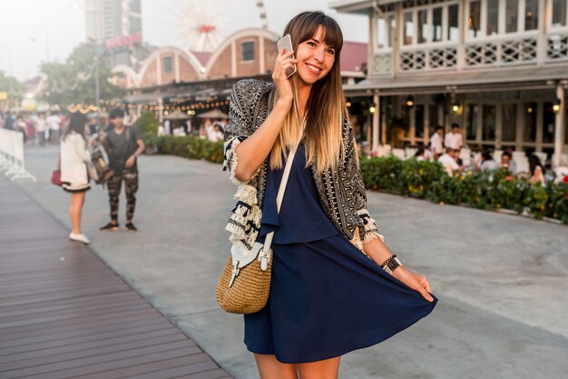 Positives Sommerporträt der fröhlichen Frau im stilvollen Outfit, das per Handy spricht und am Flussufer in Bangkok lächelt