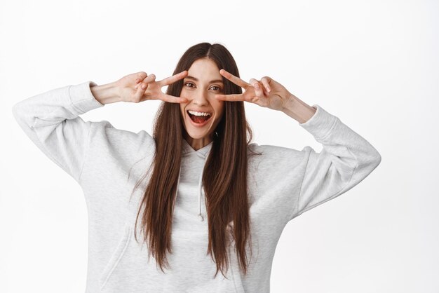 Positives Teenager-Mädchen mit langen Haaren zeigt Friedenszeichen und lächelt glücklich in die Kamera, tanzt fröhlichen weißen Hintergrund Kopierbereich