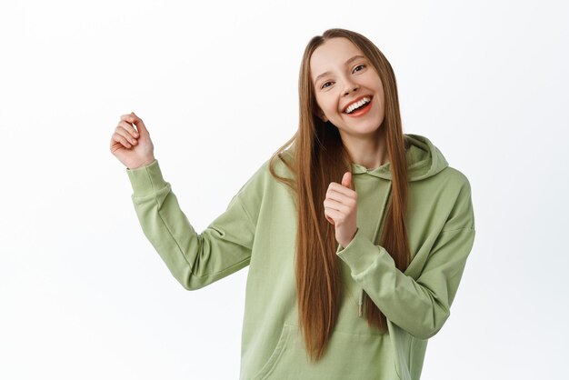 Positives Teenie-Mädchen mit langem Haar, das tanzt und Spaß hat, posiert sorglos und fröhlich im grünen Hoodie vor weißem Hintergrund