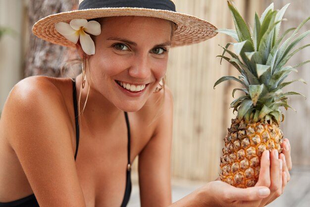 Postive entzückende Frau in Badebekleidung und Hut, genießt Sommerzeit, verbringt Urlaub im tropischen Land, hält Ananas, isst Obst, um gesund und fit auszusehen. Hübsche Frau mit exotischen leckeren Früchten