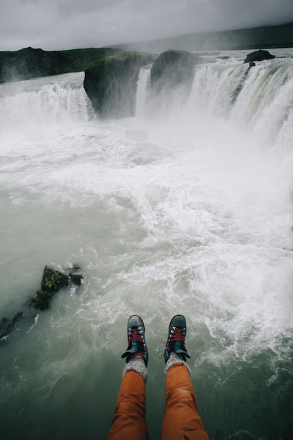 POV auf Männerbeinen in Stiefeln über Klippenwasserfall