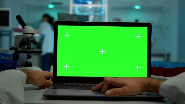 Pov-Aufnahme eines Wissenschaftlers, der am Schreibtisch sitzt und an einem Laptop mit grünem Mock-up-Bildschirm arbeitet, isolierte Anzeige. Im Hintergrund Laborforscher, die Impfstoffentwicklung untersuchen, untersuchen Proben