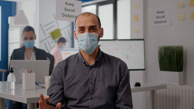 Pov-Unternehmermann mit schützender Gesichtsmaske während des Zoom-Meeting-Anrufs im neuen normalen Büro. Freiberufler, der bei einem Remote-Online-Videokonferenzanruf in die Kamera spricht