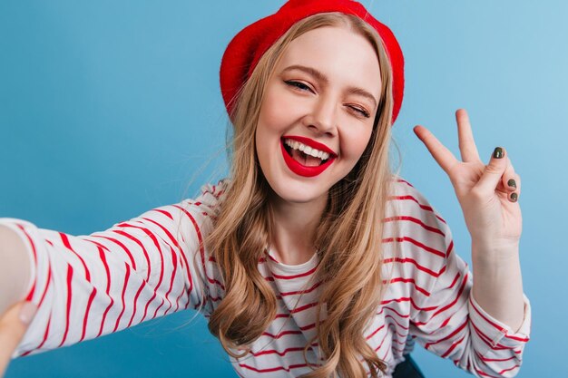 Prächtige blonde junge Frau, die Selfie auf blauem Hintergrund macht Fröhliches französisches Mädchen in Baskenmütze mit Friedenszeichen