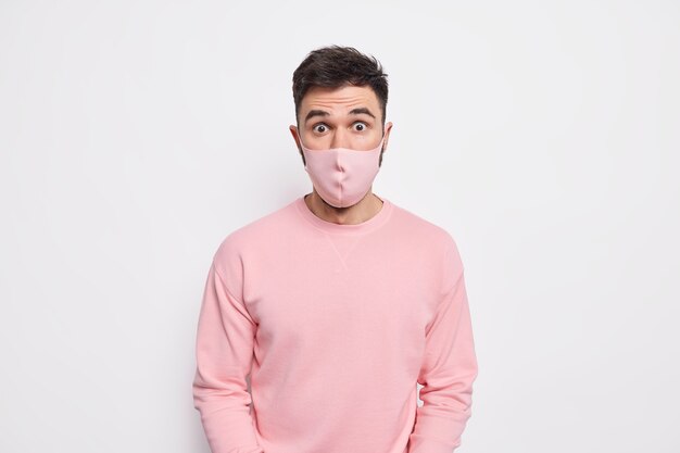 Präventions- und Sicherheitskonzept. Überraschter junger Mann trägt eine Schutzmaske im Gesicht verhindert die Ausbreitung des Coronavirus und erfährt schockierende Statistiken in einem rosa Pullover pink