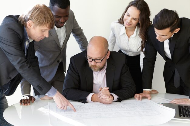 Produktive Teamarbeit im Büro. Der Hauptingenieur für Brillen hört neuen Ideen seines Kollegen zu. Kaukasischer Mann, der Finger auf schematische Zeichnungen zeigt. Mitarbeiter, die sein Angebot mit einem Lächeln genehmigen.