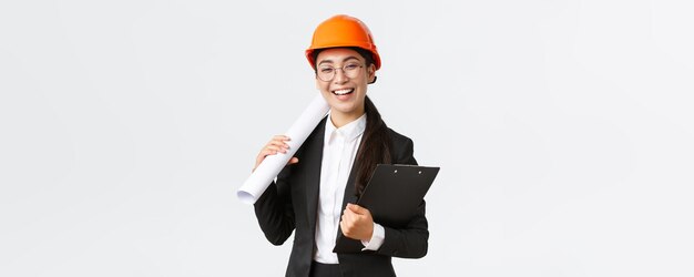 Professionelle glückliche asiatische Architektin Bauingenieurin in Helm und Business-Anzug mit Bauplänen und Klemmbrett mit lächelndem fröhlichem weißem Hintergrund der Baudokumentation