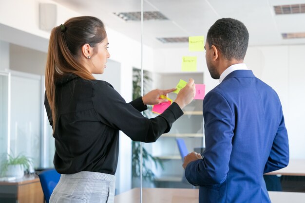 Professionelle junge Kollegen, die mit Aufklebern arbeiten. Fokussierte Arbeiterin, die Notiz auf Glas klebt. Konzentrierter Geschäftsmann im blauen Anzug, der neben ihr steht. Konzept für Teamarbeit, Geschäft und Zusammenarbeit