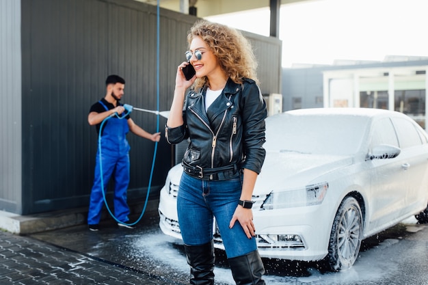 Professionelle Waschmaschine in blauer Uniform, die Luxusauto mit Wasserpistole auf einer Open-Air-Autowaschanlage wäscht