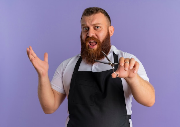 Professioneller bärtiger Friseurmann in der Schürze, die den Arm der Schere hält, der verwirrt und unzufrieden steht, der über lila Wand steht