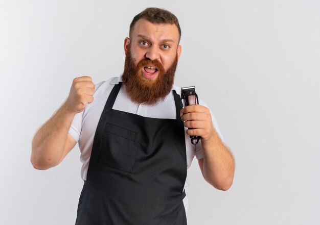 Professioneller bärtiger Friseurmann in der Schürze, die Haarschneidemaschine hält, die Faust mit dem verärgerten Gesicht zusammenhält, das frustriert über weißer Wand steht